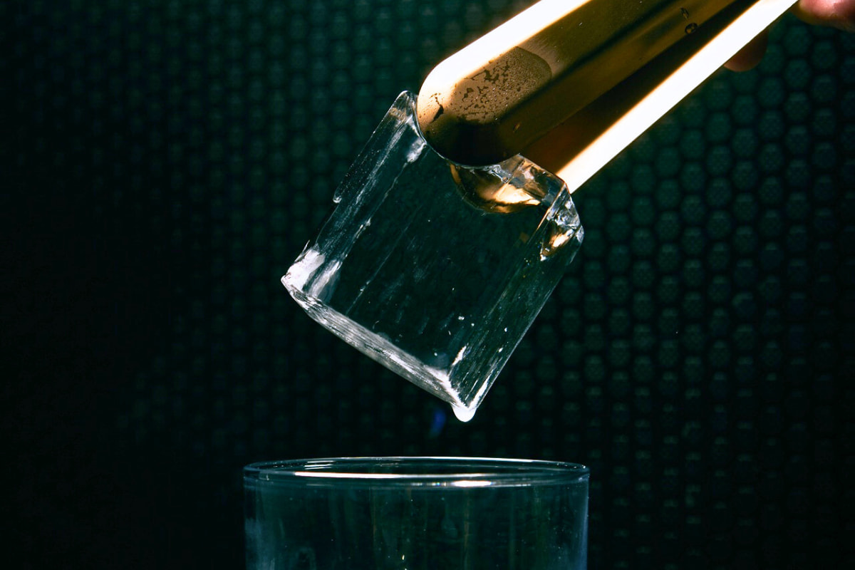 Cubito de hielo gourmet transparente cogido con pinzas para ser introducido dentro de un vaso de cristal